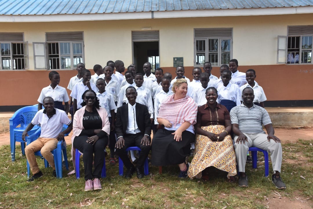 Poetry for Peace Club pupils standing behind the seated PJF team outside a classroom at Kweyo Primary School